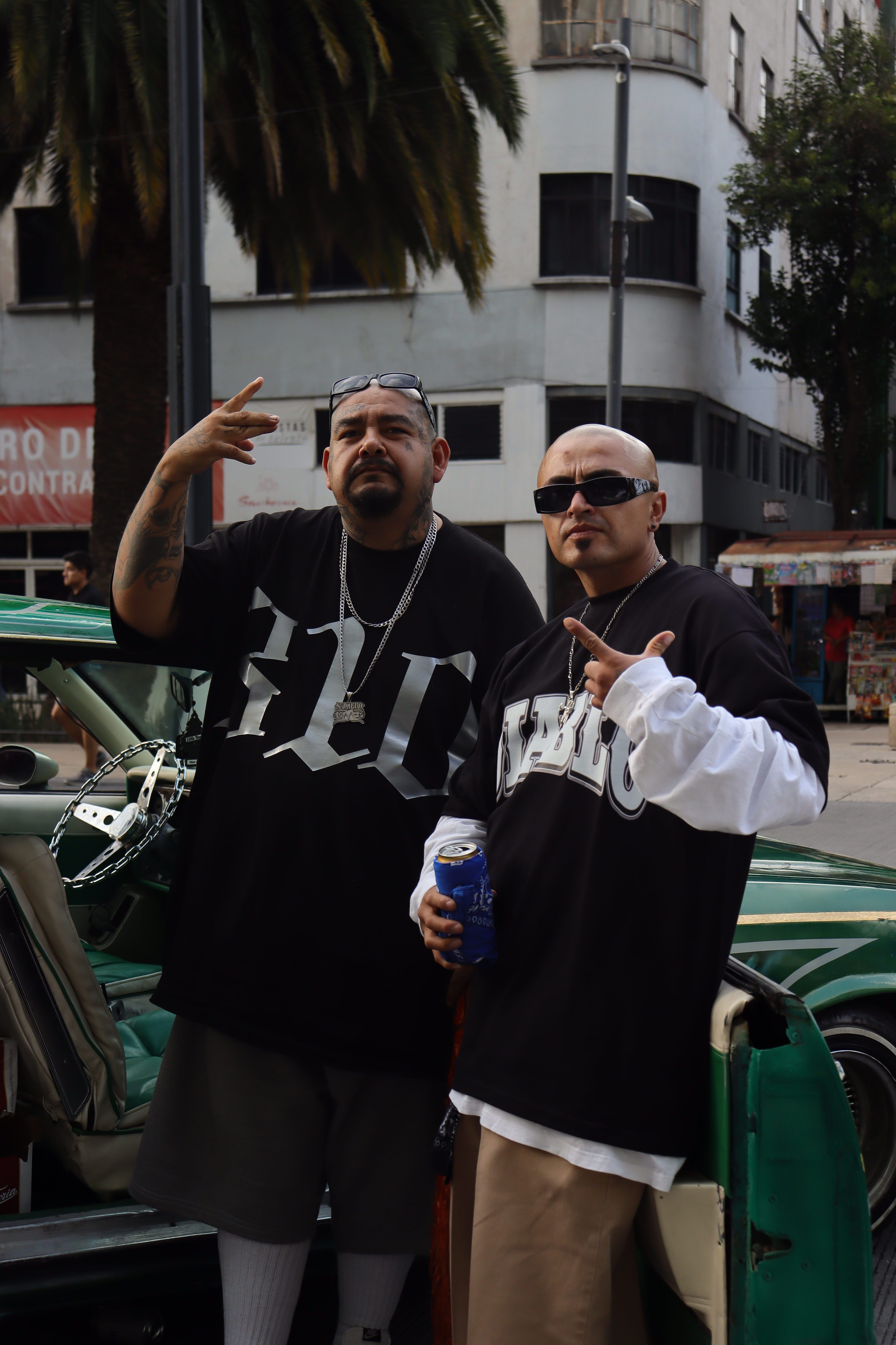 Two men posing confidently in streetwear beside a green car in a city setting.