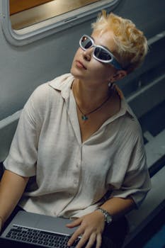 Fashionable woman with short blonde hair wearing sunglasses and holding a laptop indoors.