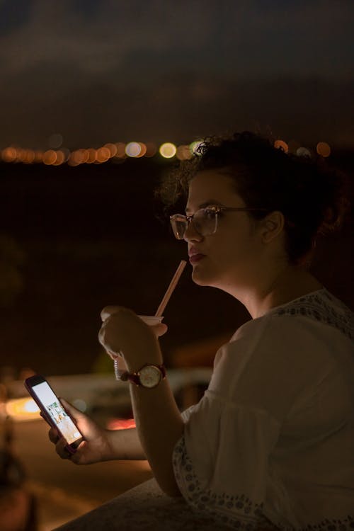 Woman Using Smartphone and Holding Plastic Cup With Straw Outdoors at Nighttime