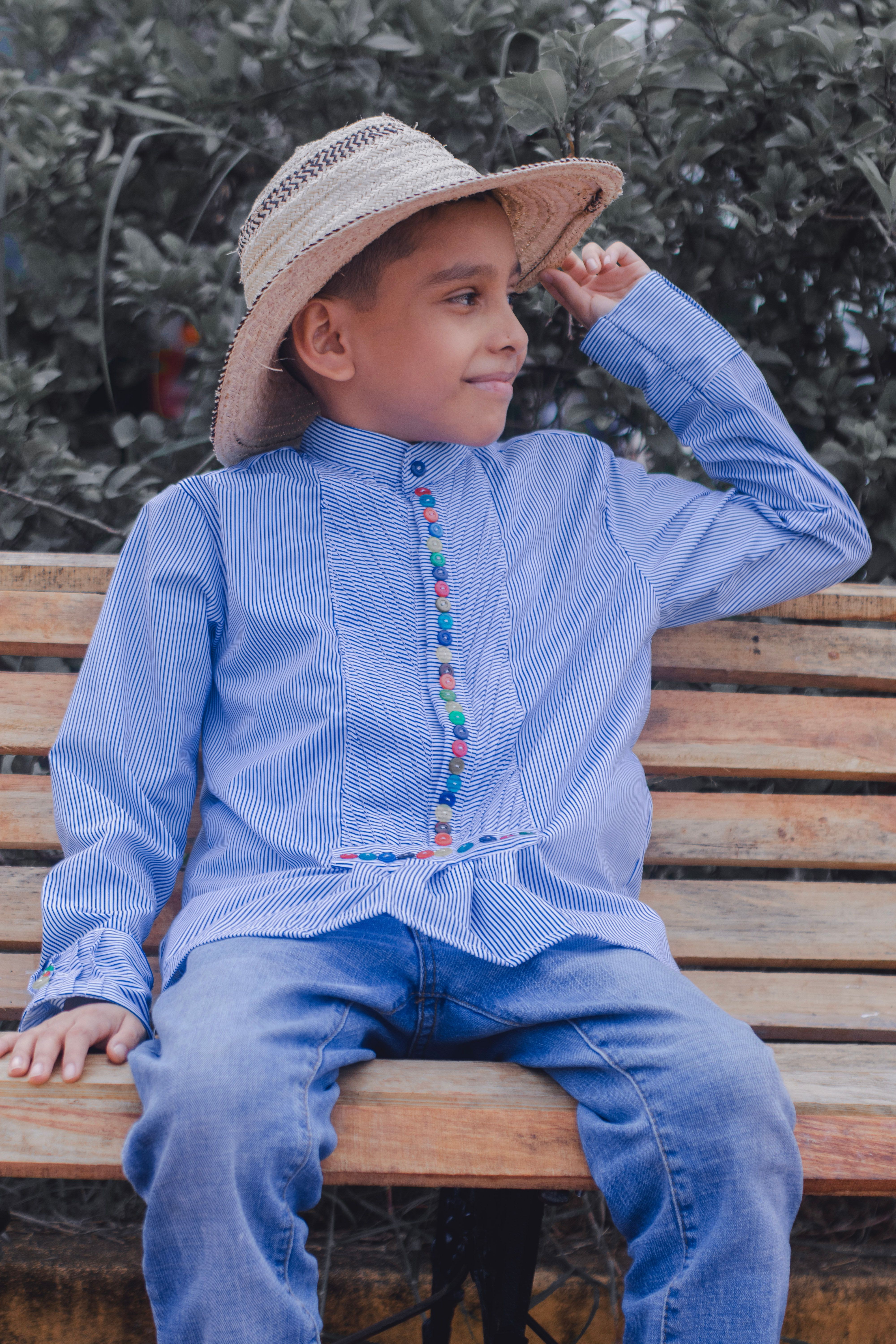 Fotomural Un niño joven que lleva puesta una camisa azul elegante y vaqueros se sienta en un banco con un sombrero de paja al aire libre.