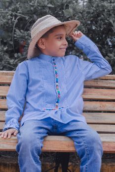 A young boy wearing a stylish blue shirt and jeans sits on a bench with a straw hat outdoors.
