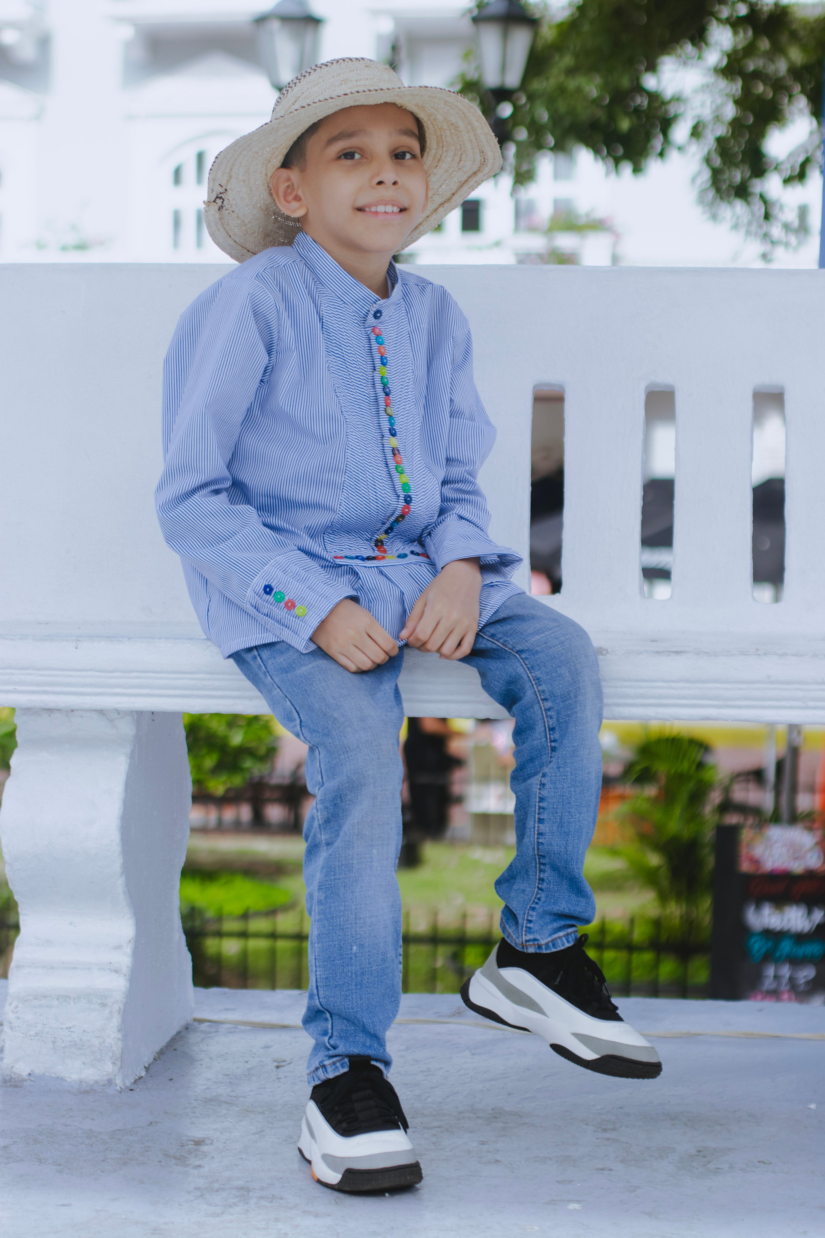 Um menino sorridente em uma camisa azul e jeans sentado em um banco branco ao ar livre.