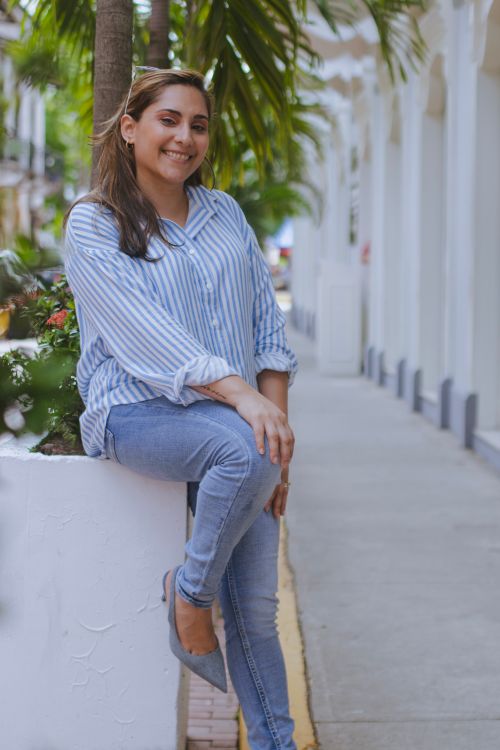 Free Woman in casual attire smiling while sitting on a planter in a lush, urban street setting. Stock Photo