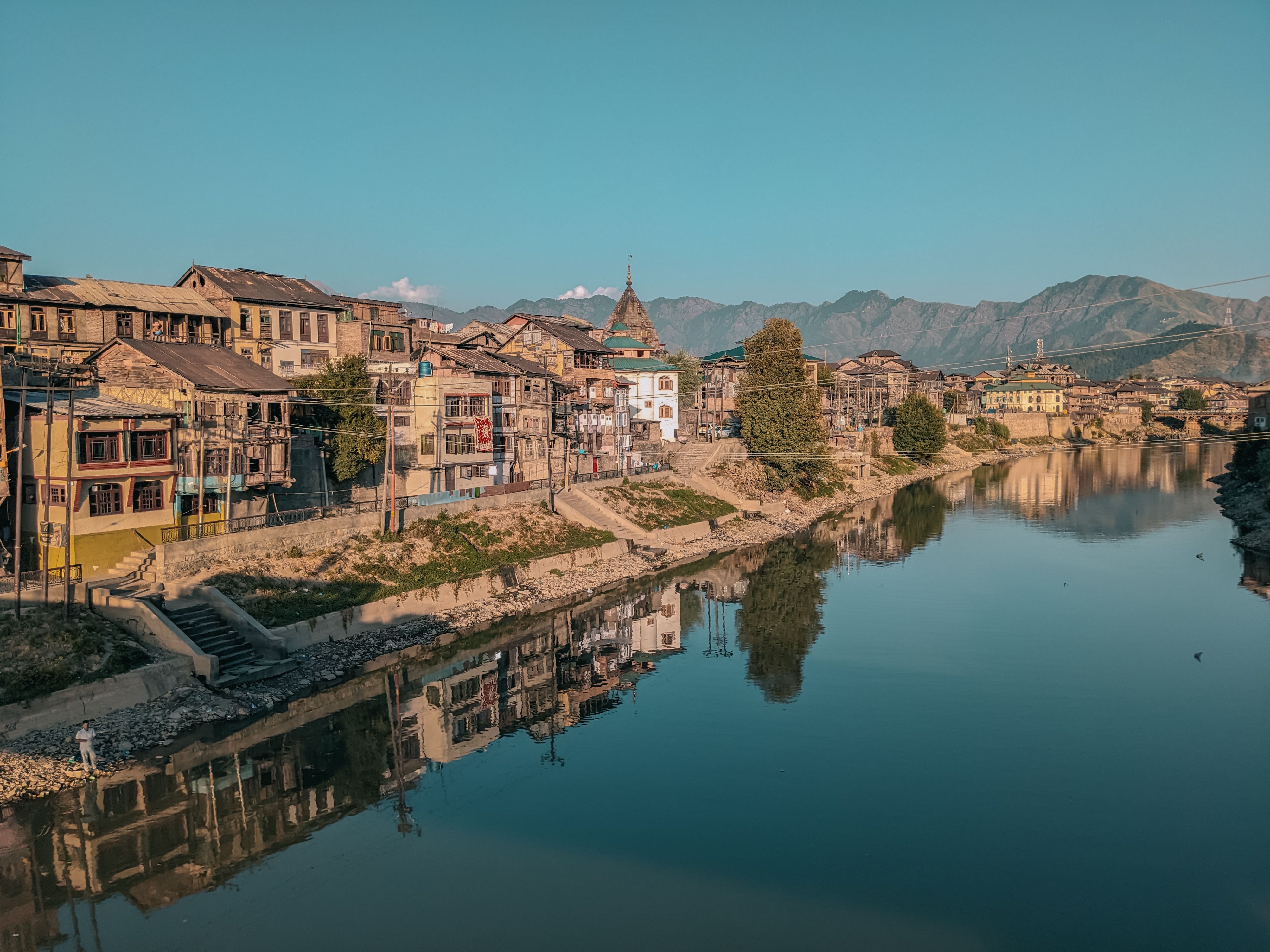 Atmospheric view of the Jhelum River in Srinagar, India, showing the riverfront setting around Sher Garhi Palace and traditional buildings against a mountain backdrop.