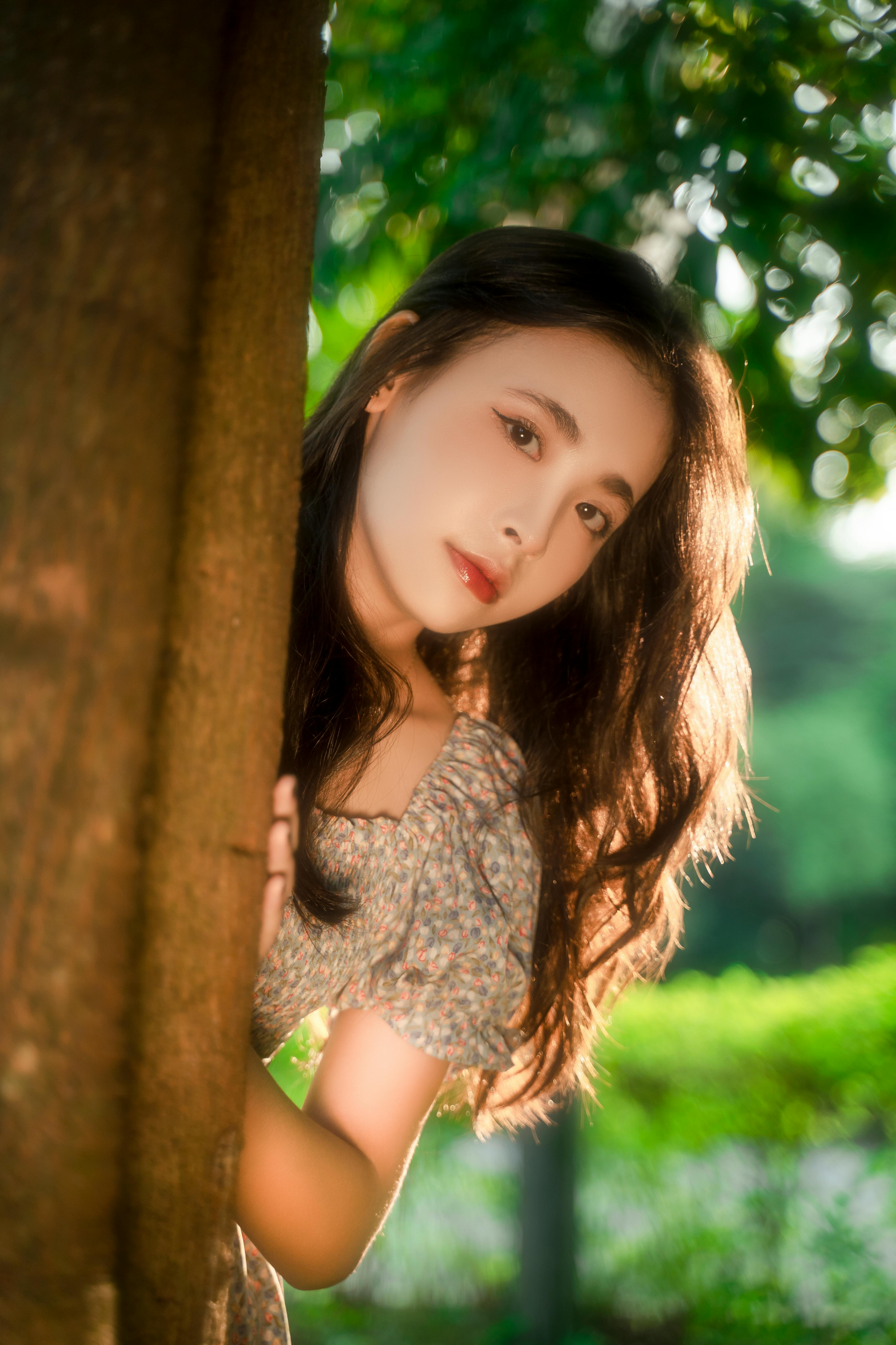 A young woman with long hair embraces the warm sunlight while leaning against a tree in a vibrant park setting.