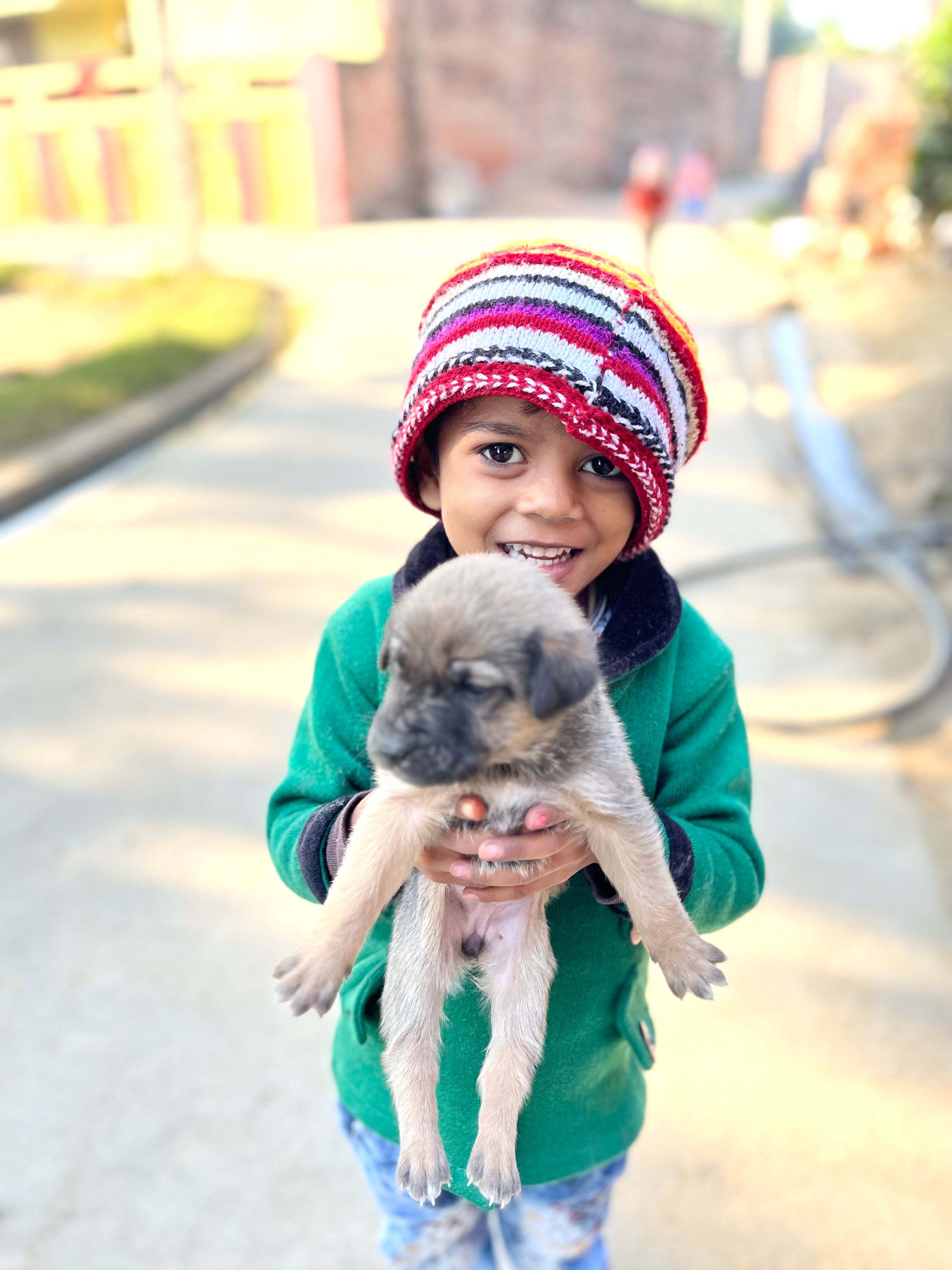 A joyful child wearing a colorful beanie holds a puppy on a sunny street, showcasing happiness and innocence.