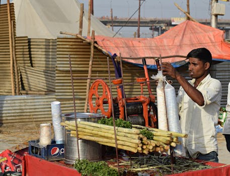 A man prepares fresh sugarcane juice at a street stall in a village setting using a manual juicer.