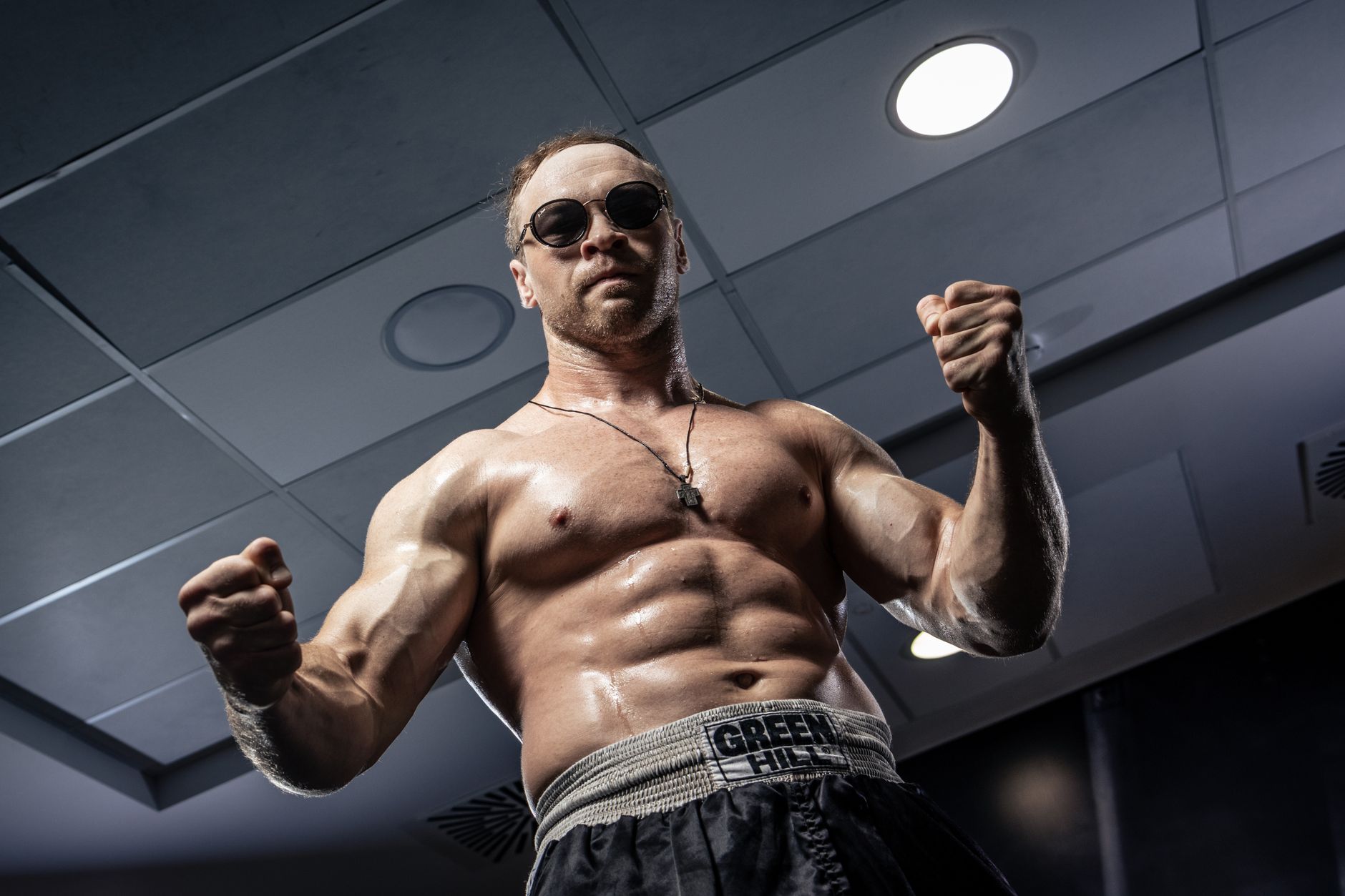 A strong, shirtless man in sunglasses posing confidently in a gym under ceiling lights.