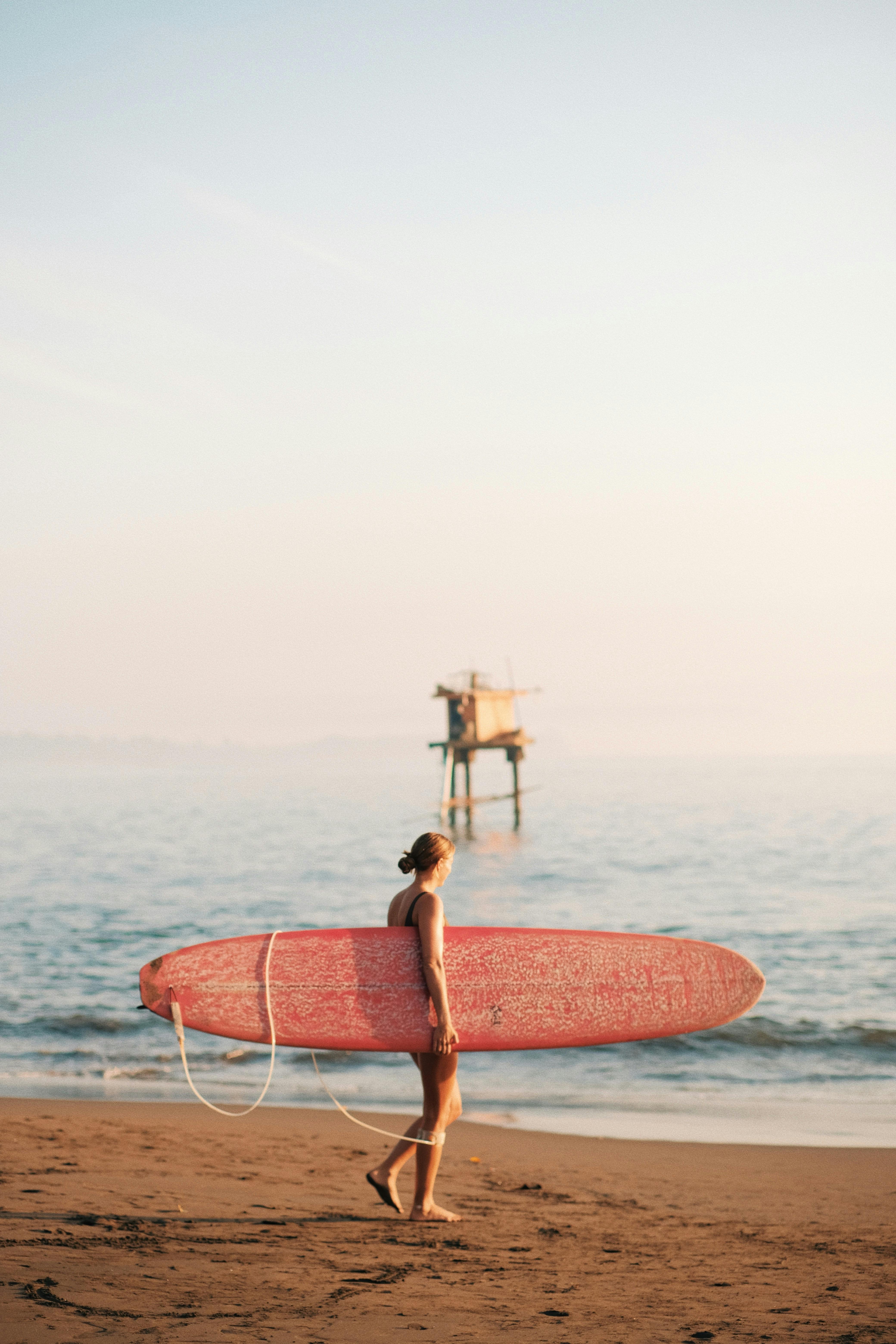 A woman carrying a surfboard walks along a serene beach during a summer sunset.