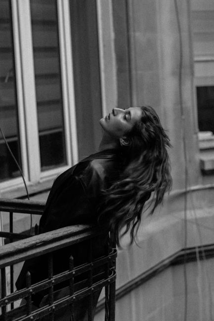 Black and white portrait of a woman leaning on a city balcony, capturing a moment of introspection.