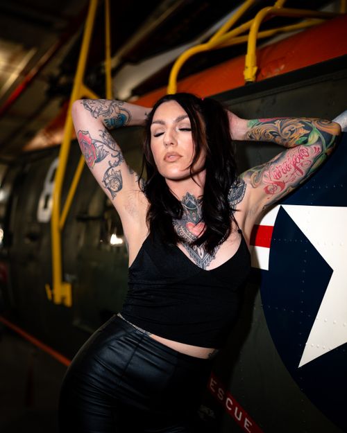 Free A tattooed model in a black outfit poses confidently by an aircraft indoors. Stock Photo