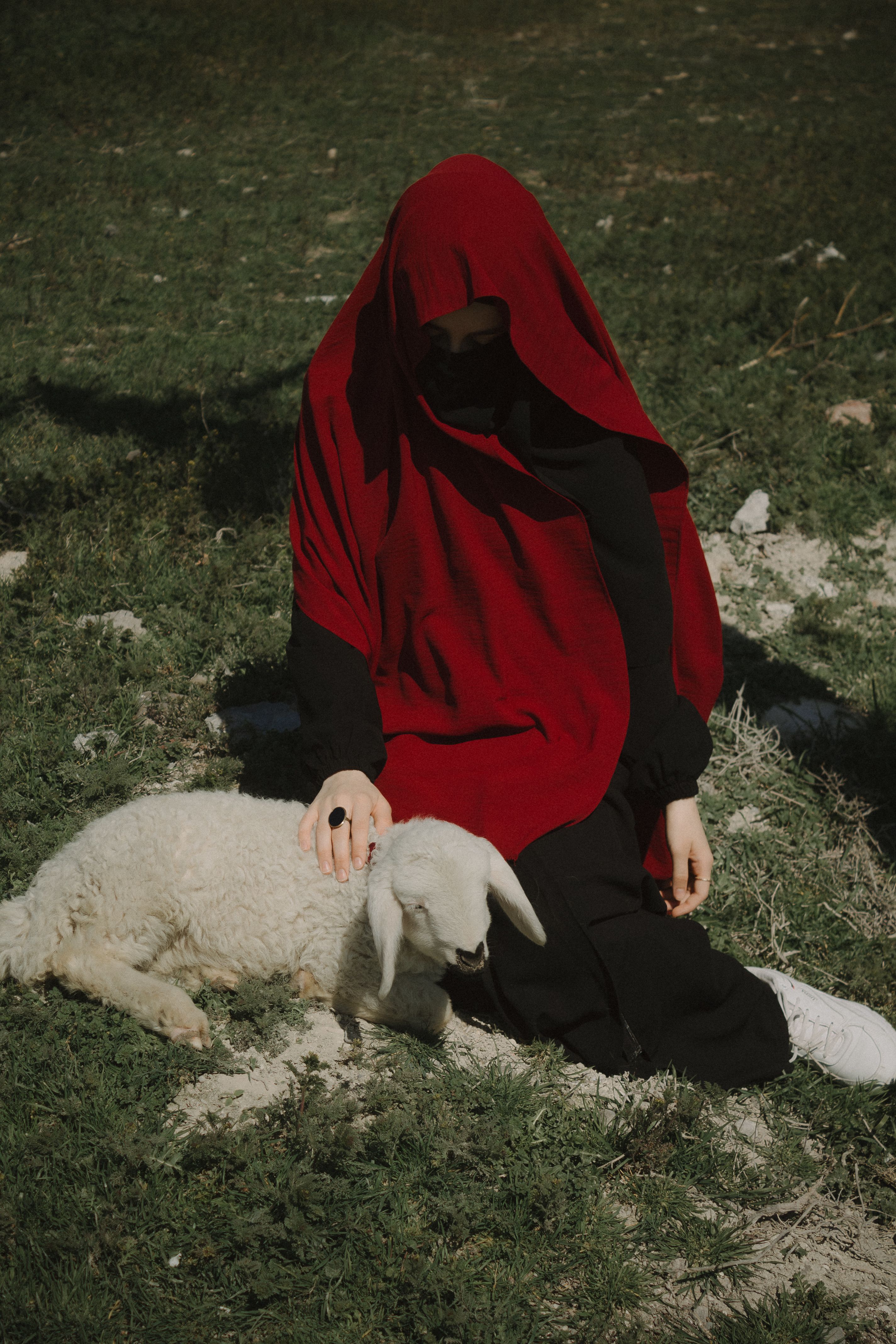 A woman in a red veil gently strokes a lamb in a grassy pasture, representing tranquility and tradition.