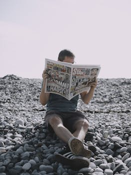 Man enjoying leisure time reading a newspaper on a stony beach under bright daylight.