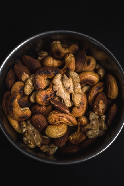 Close-up of assorted roasted nuts in a steel bowl on a black background, showcasing almonds, walnuts, and cashews. - buying dry fruits online Pakistan