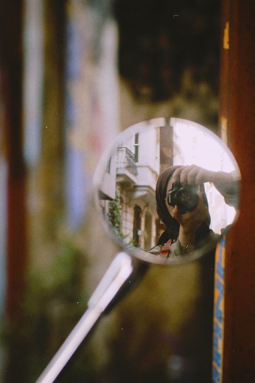 Free A woman's reflection while taking a photo in a motorbike mirror in an urban setting. Stock Photo