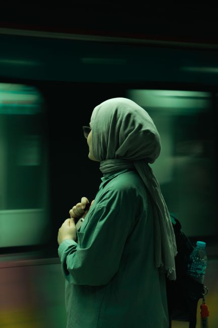 Side view of a woman wearing a headscarf waiting on a subway platform with a backpack.