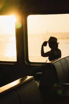 A silhouette of a person wearing a hat on a ferry at sunset, with glowing sunlight through windows.