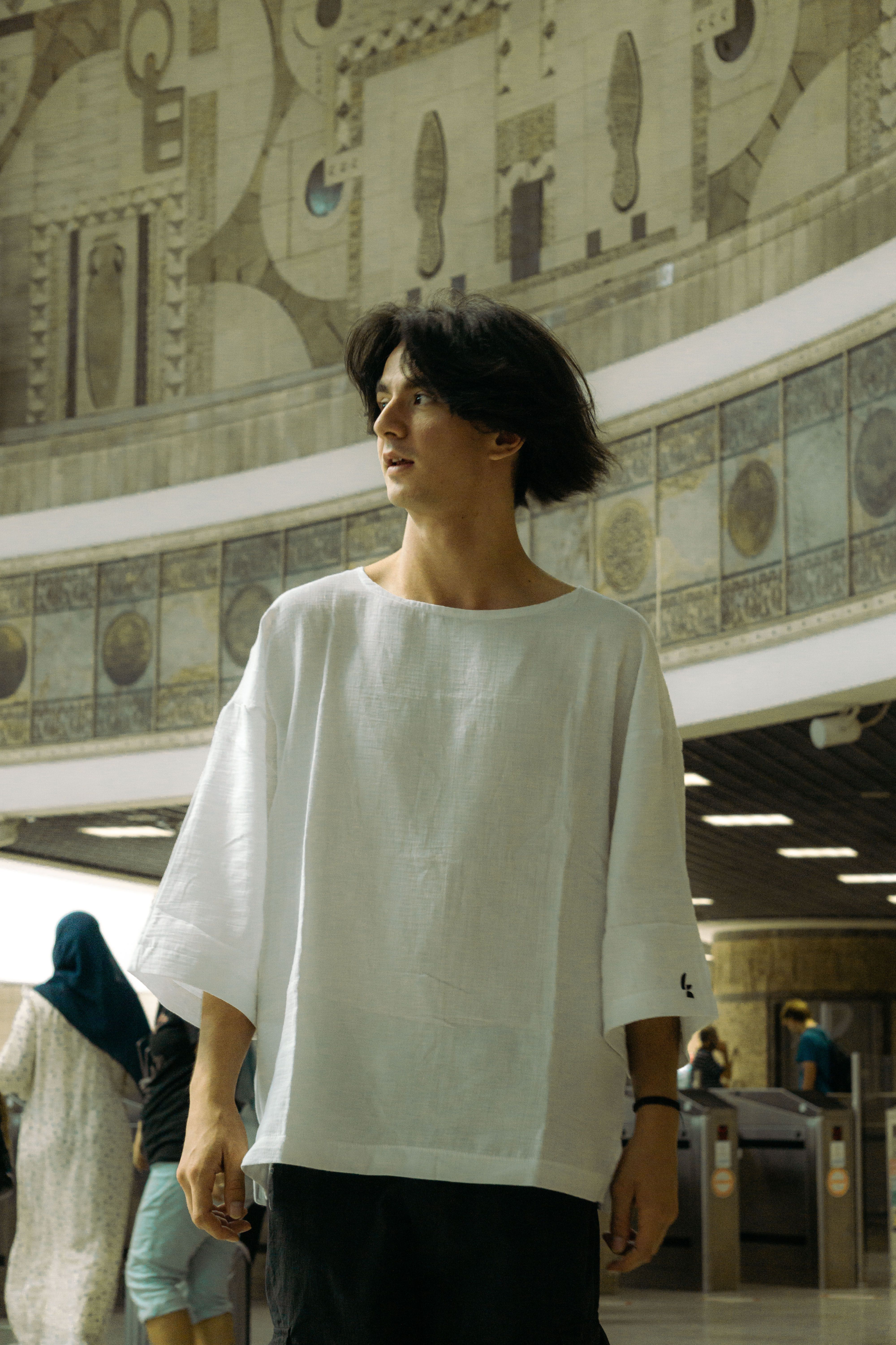 A stylish young adult in an oversized white t-shirt stands in an urban environment with intricate architectural details.