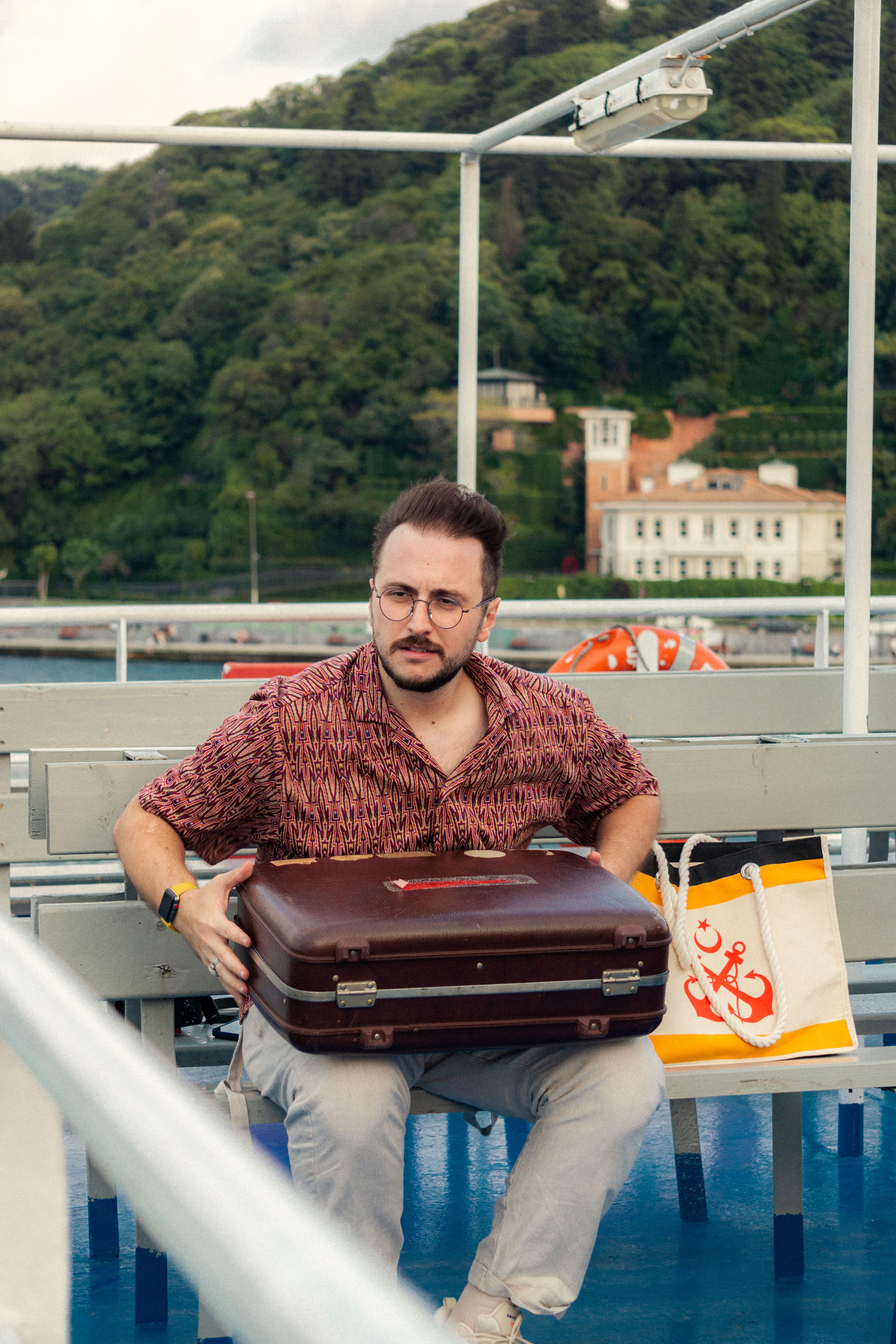 A middle-aged man sitting on a ferry, holding luggage, surrounded by scenic landscape.
