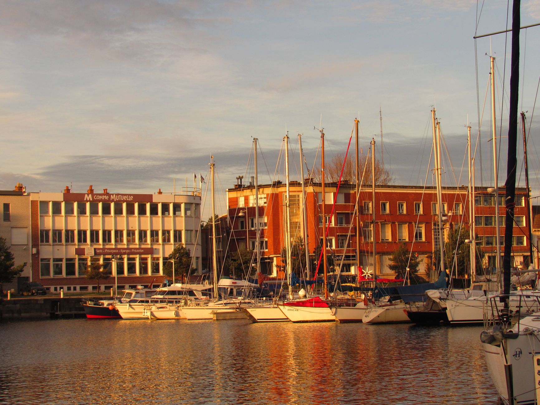 Sailboats moored in Calais harbor with colorful buildings at sunset.