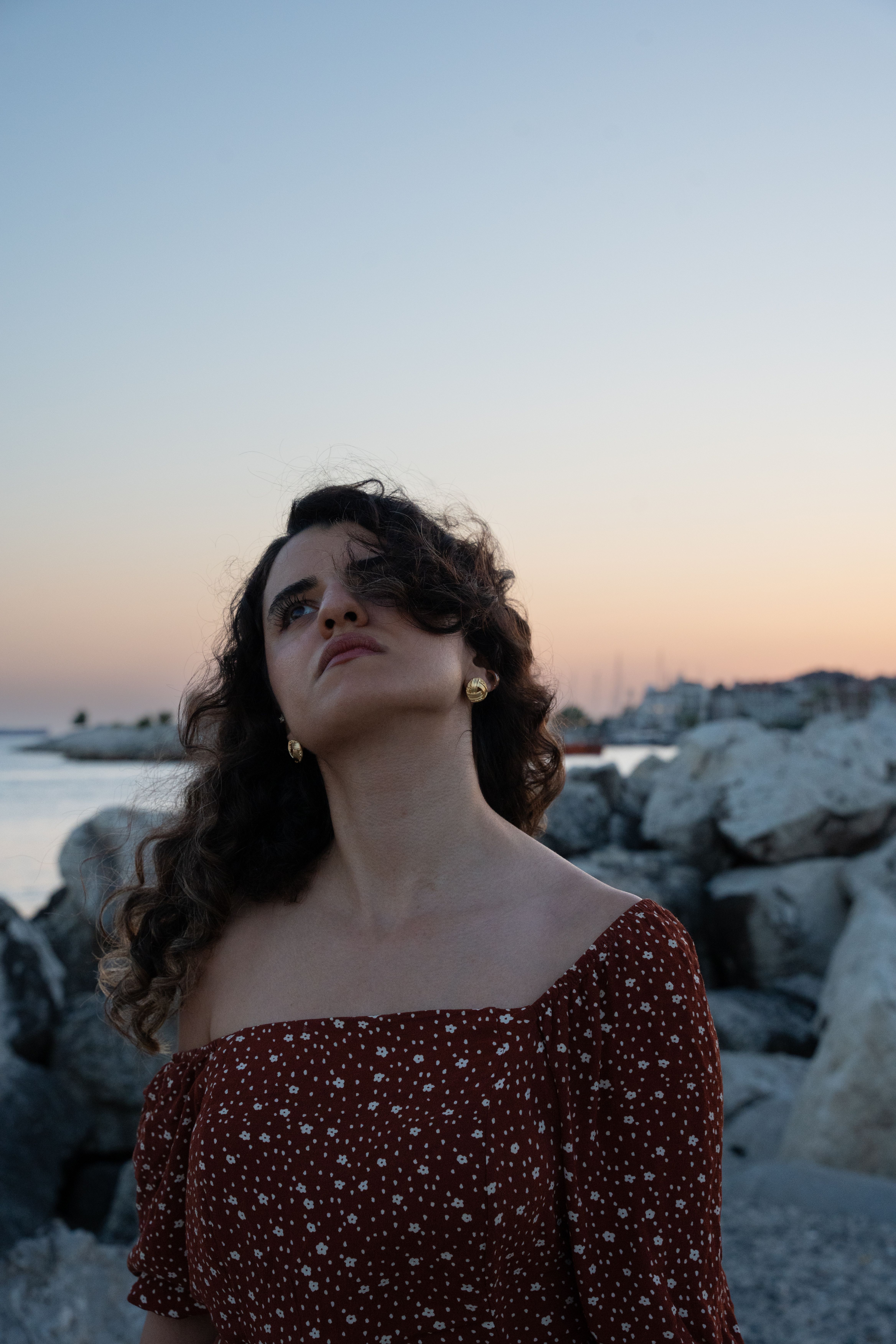 Young woman in floral dress at rocky beach during sunset, capturing a serene moment.