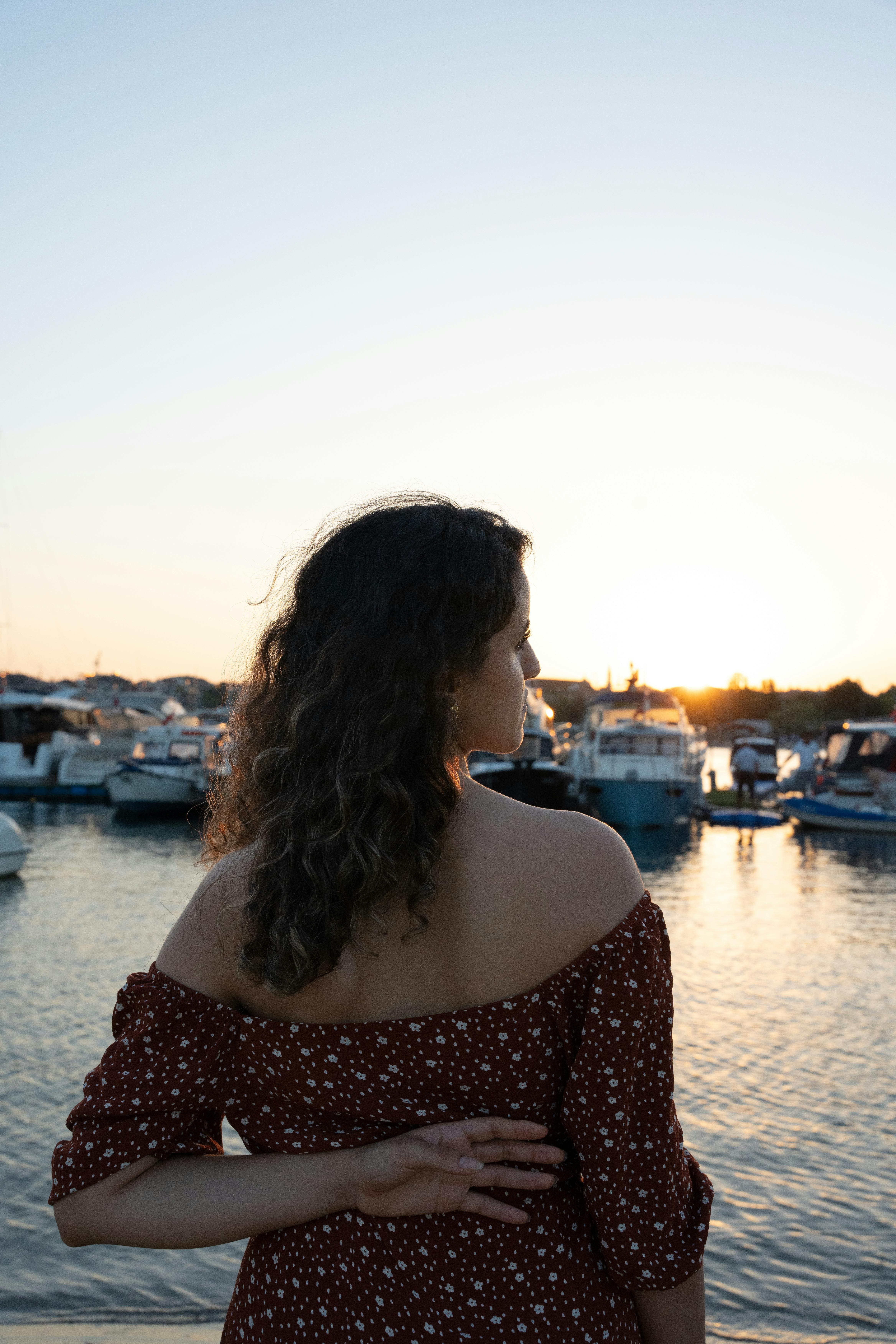 A woman in a red dress stands by the marina, admiring the boats at sunset.