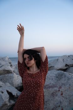A woman in a floral sundress poses gracefully on a rocky shoreline at twilight.