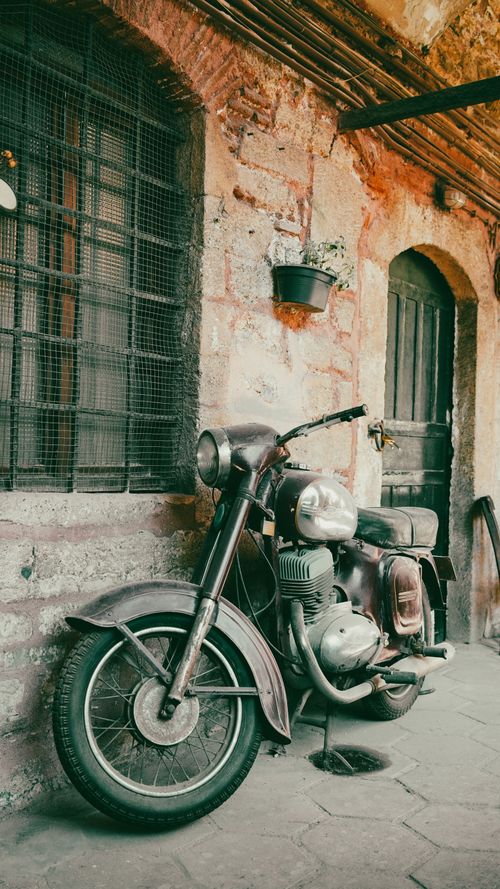 Free A classic motorcycle is parked on a charming, vintage street beside a stone wall. Stock Photo