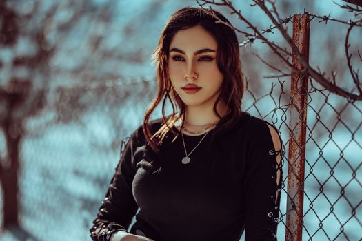 Young woman standing by a fence in winter, showcasing fashion and beauty in a cold setting.