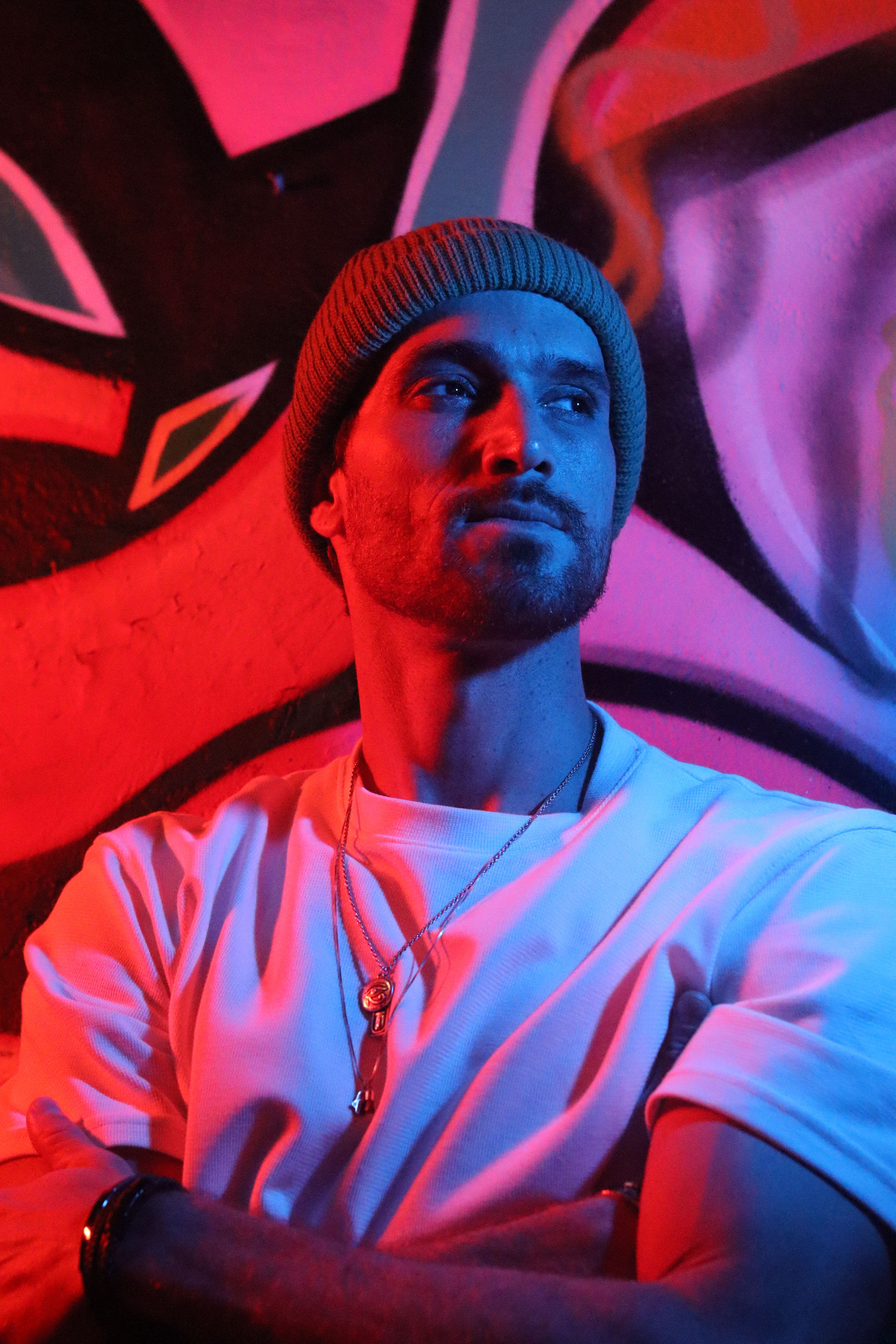 Stylish portrait of a man in neon lighting with graffiti background, wearing beanie and necklace.