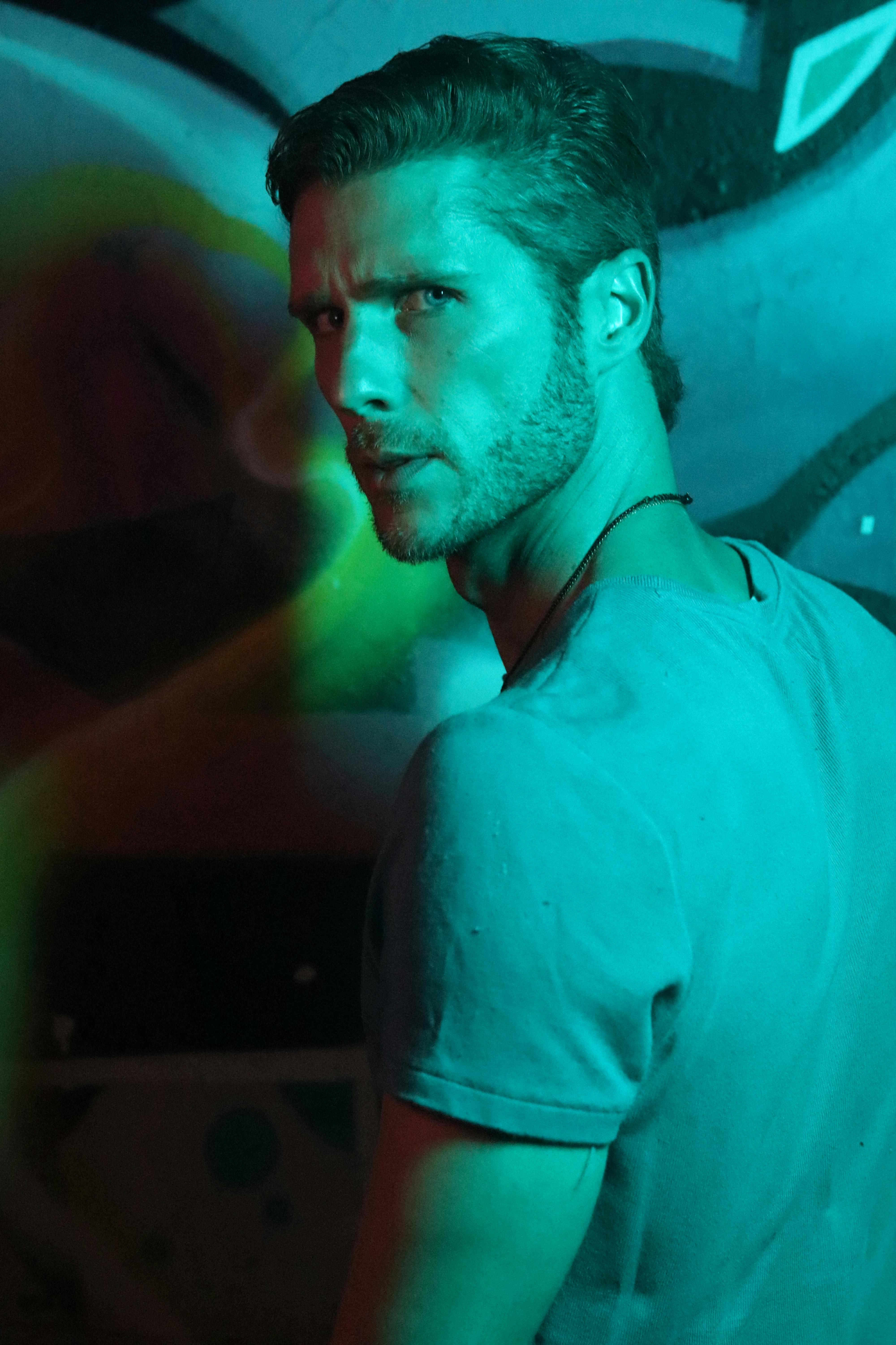 A man with short hair stands in an urban area, back view illuminated by neon lights.