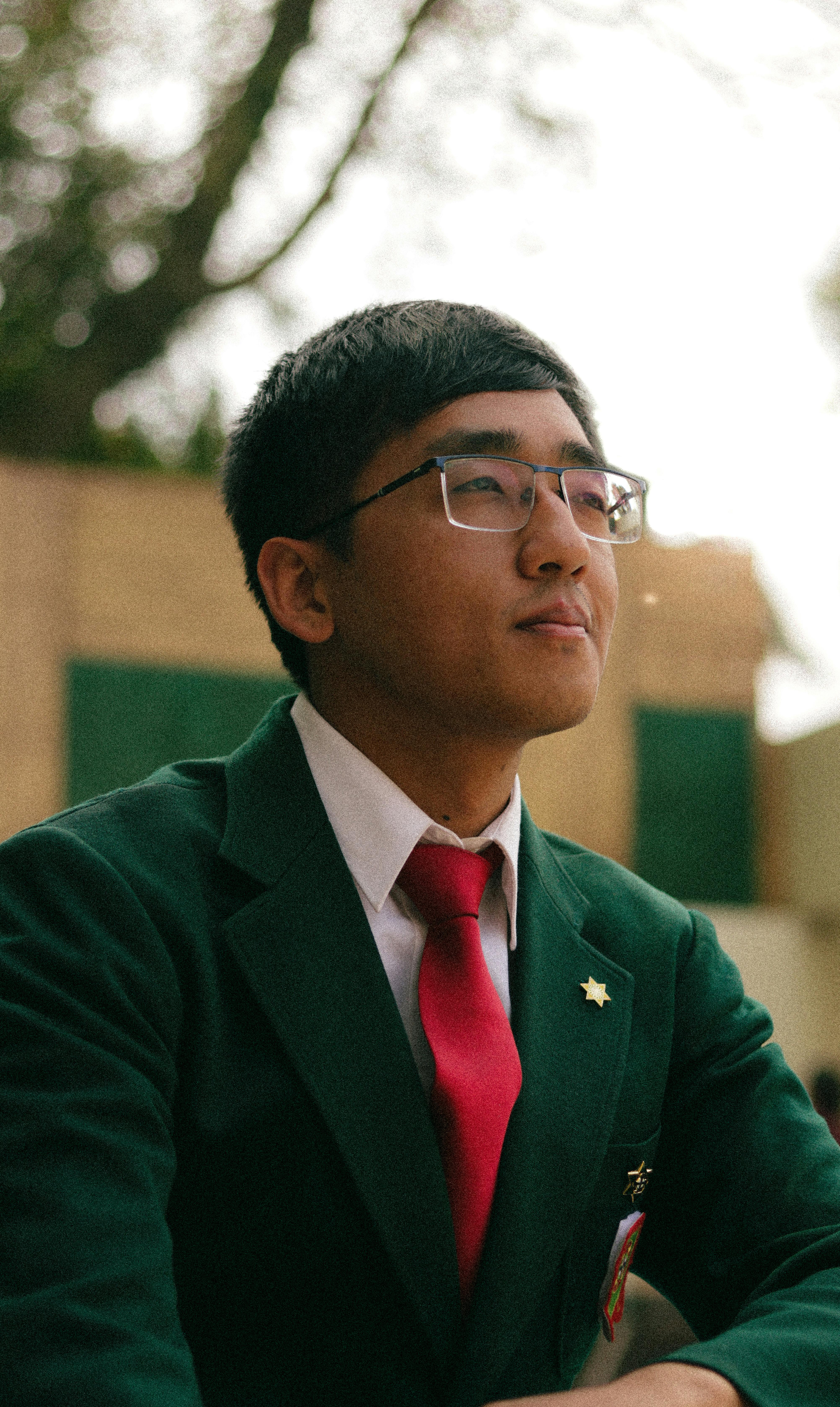 Portrait of a young Asian man in formal attire wearing a green blazer and red tie, sitting outdoors.