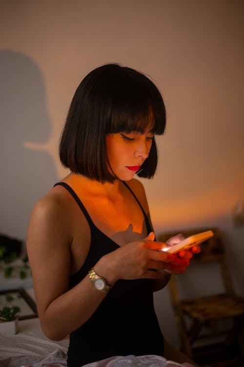 Woman in a Black Bodysuit Sitting on a Bed Using Smartphone