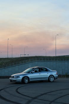 A sleek silver car parked on an open road during sunrise under soft skies and street lamps.