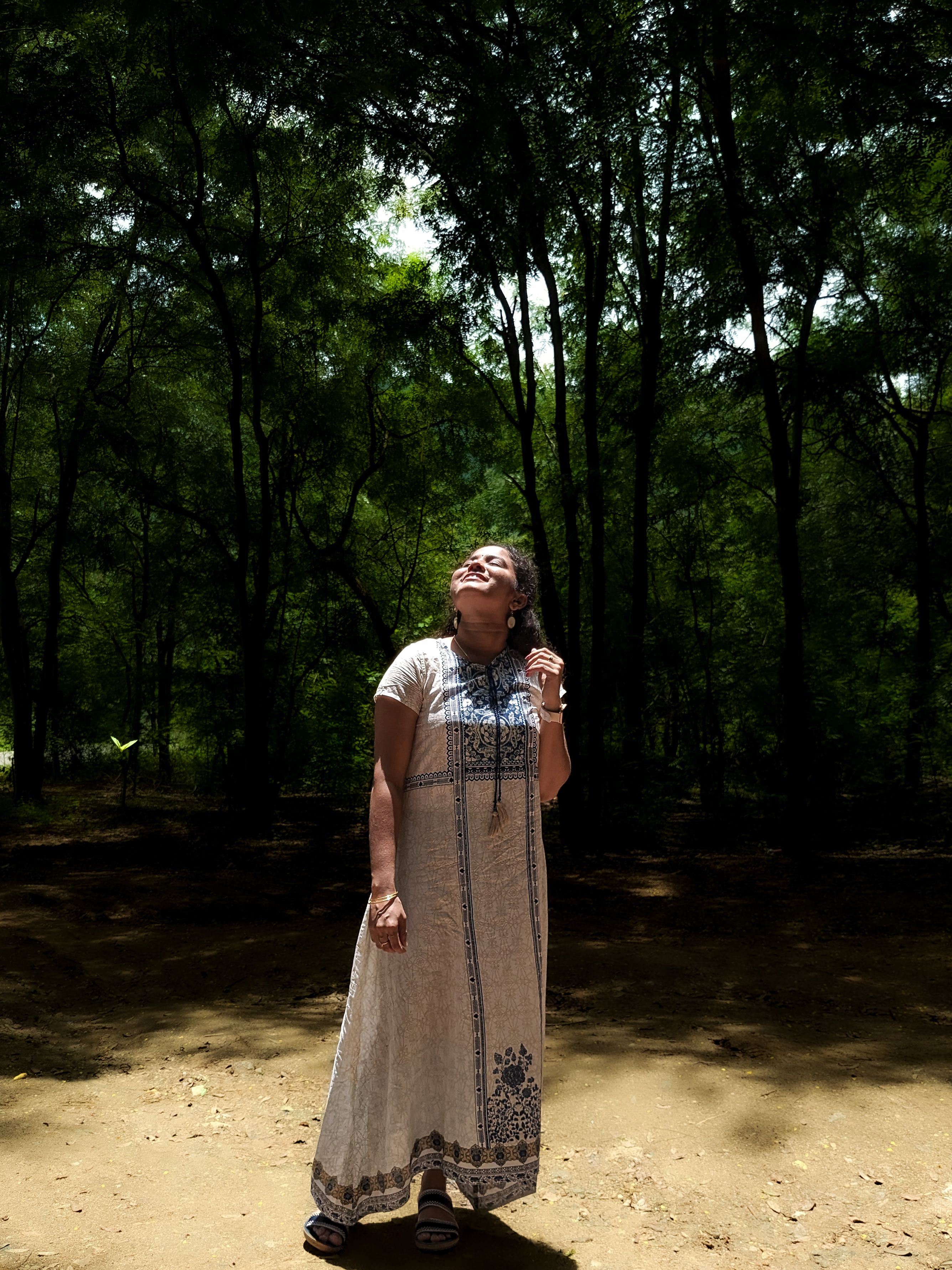 Woman in a maxi dress standing in a sunlit forest clearing, embracing nature.