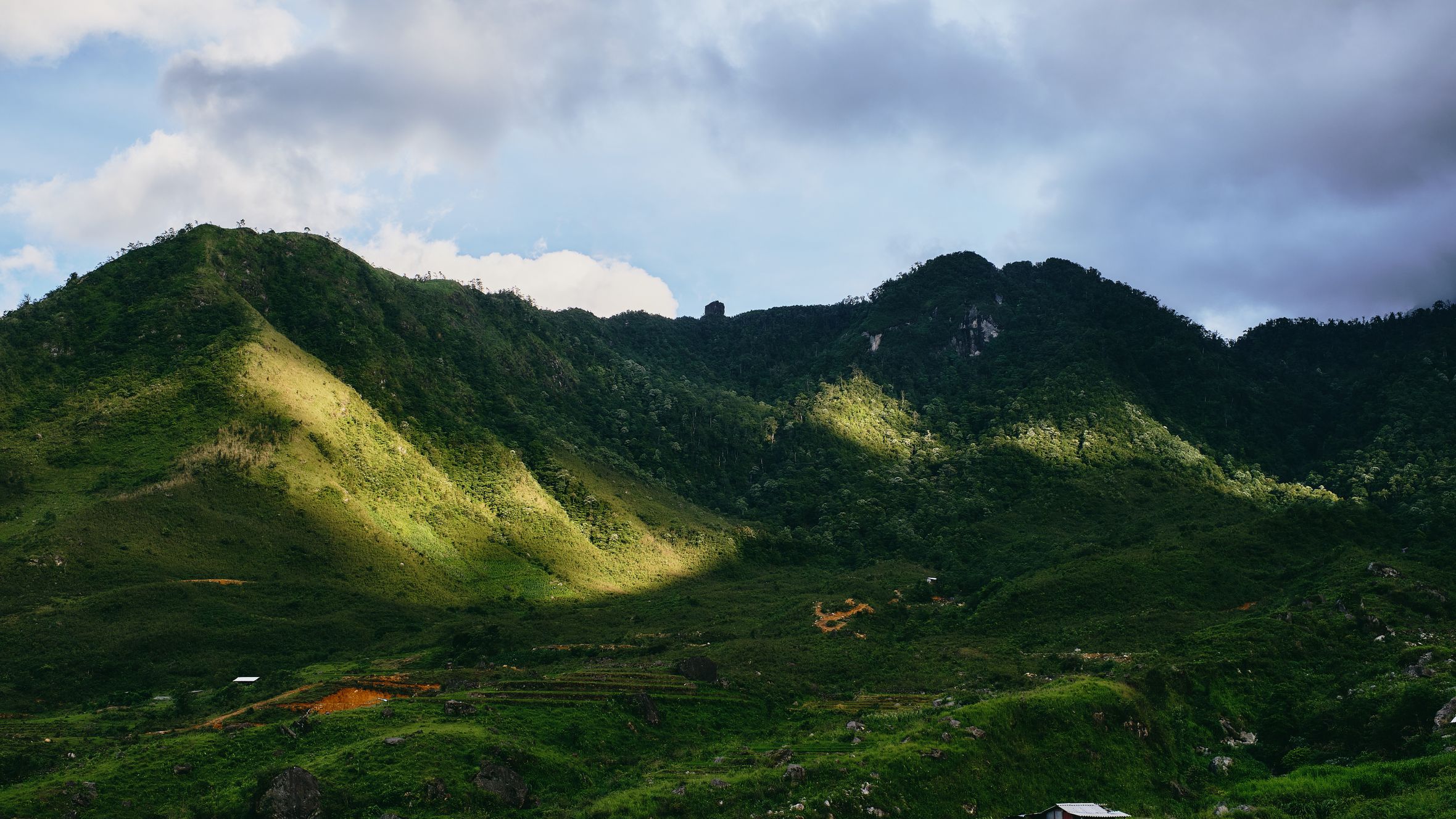 Breathtaking aerial view of lush green hills and valleys in Lào Cai, Vietnam, under a cloudy blue sky.