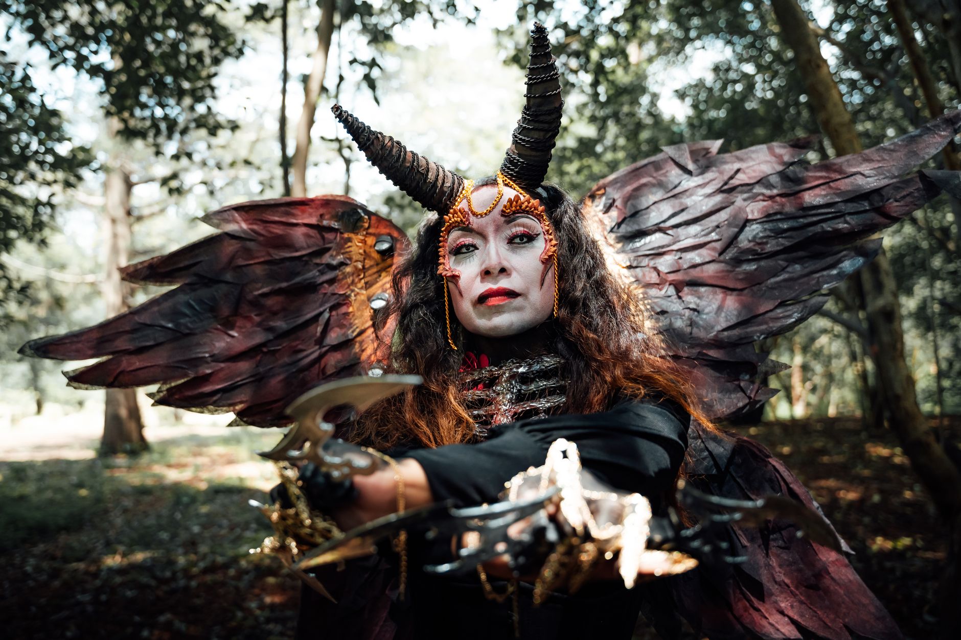 A woman in artistic costume poses with horns and wings in a forest setting.