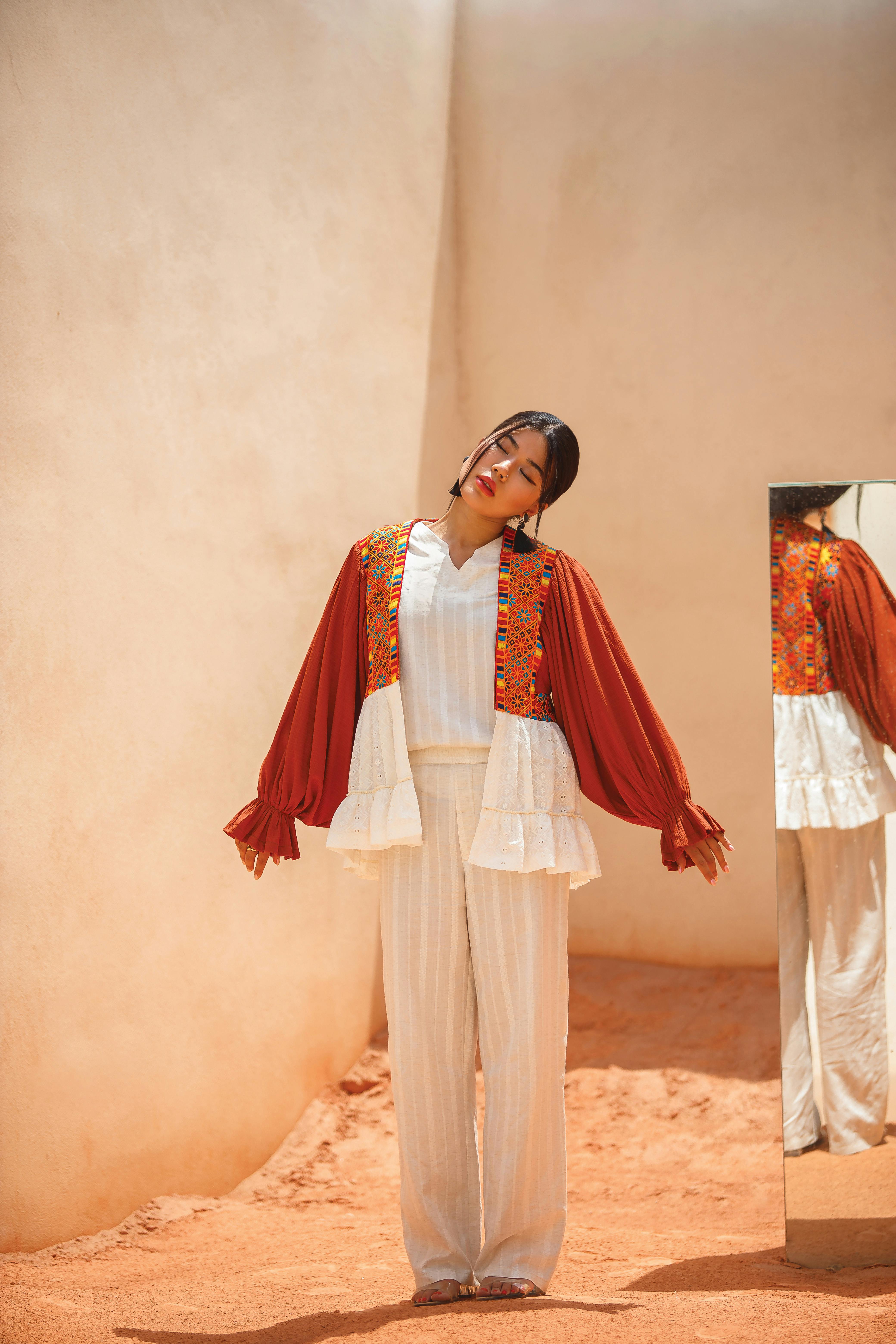 Elegant woman in bohemian outfit poses by mirror in a desert setting, reflecting contemporary style.