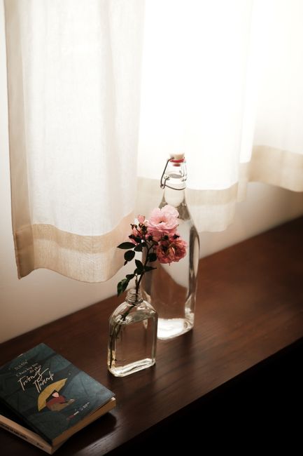 A serene still life featuring pink flowers in a bottle by a curtain-lit window.