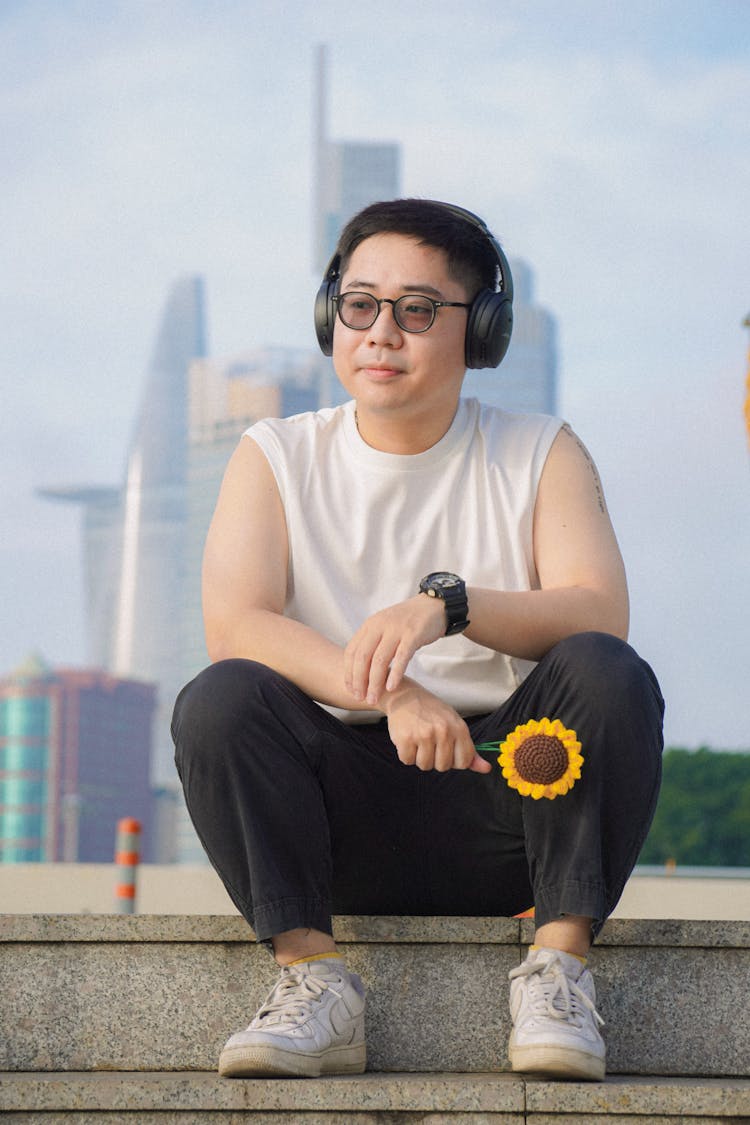 Young Man Wearing Headphones And Holding A Sunflower Sitting On Steps In City 