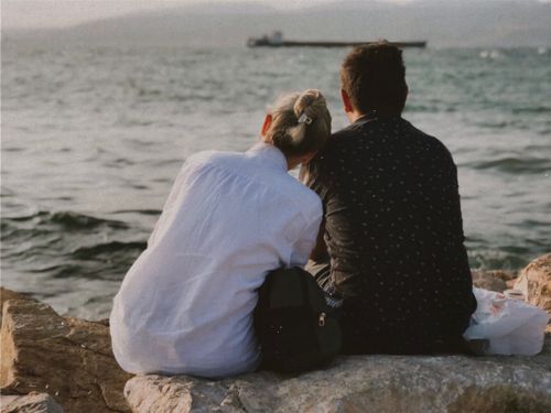 Free Couple sitting on rocks by the sea, enjoying a peaceful sunset moment. Stock Photo