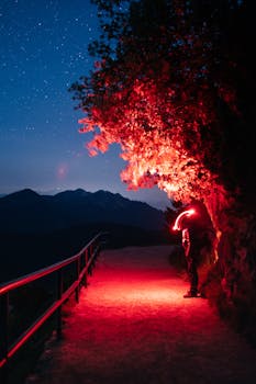 A lone hiker with a red light walking on a mountain path under a starry night sky.