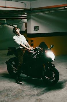 A young man leans confidently on a motorcycle in a dimly lit parking garage.