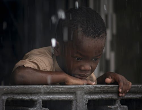 Free A young boy gazes out from a balcony during rain, evoking emotions of solitude. Stock Photo