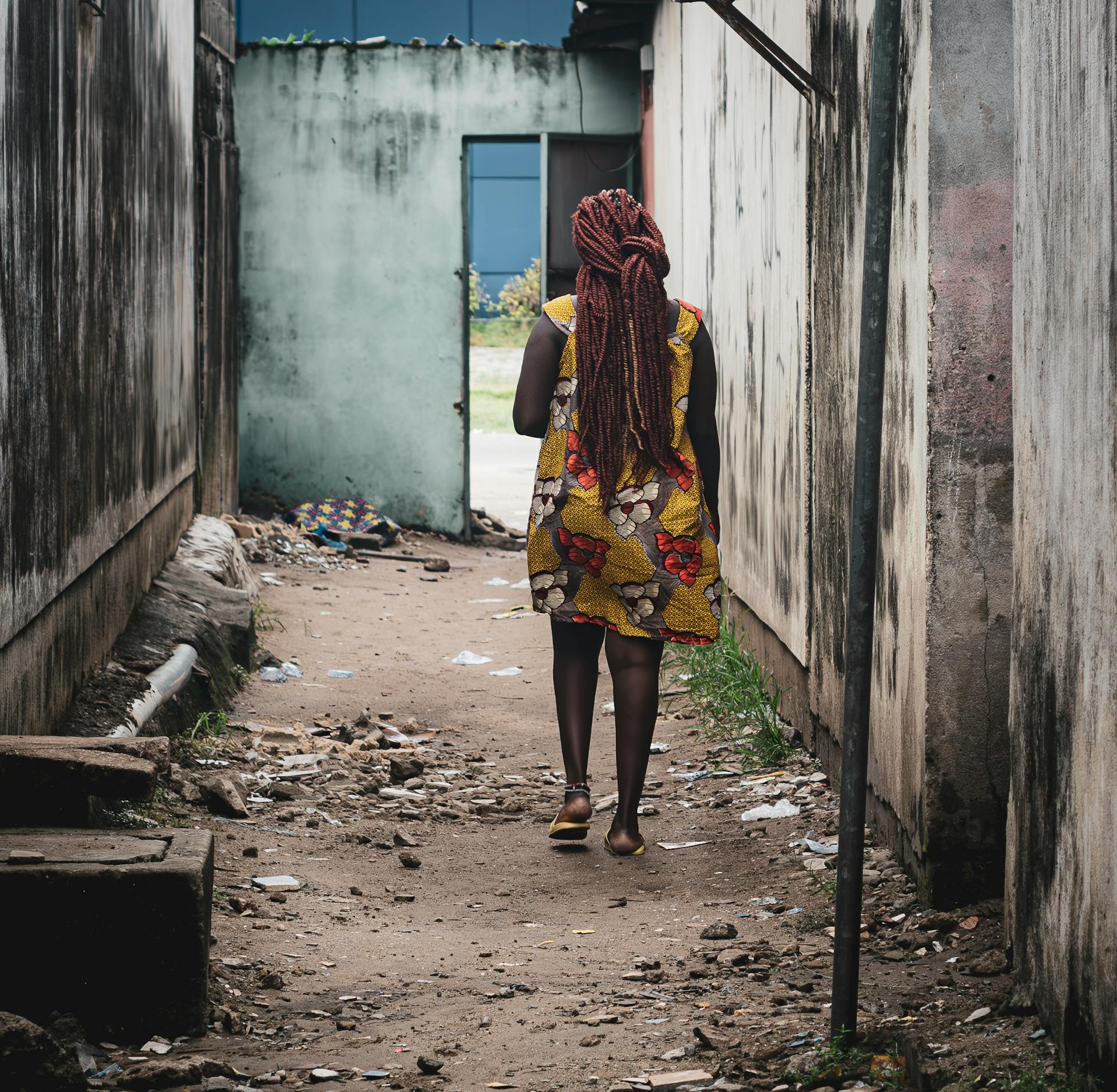 African woman with dreadlocks walking through a narrow alley in an urban setting.