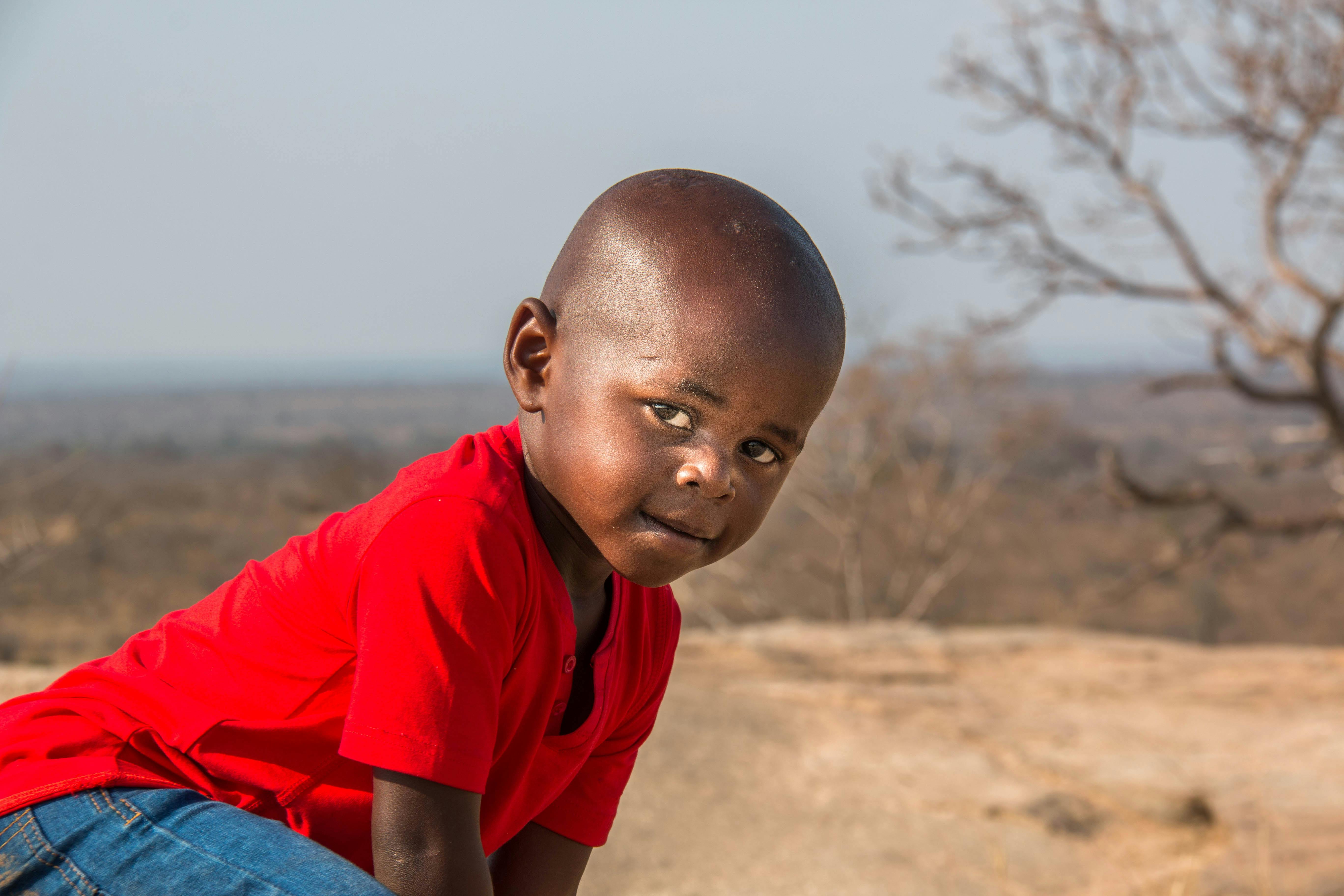 A young boy crouches outdoors in South Africa, wearing a red t-shirt and jeans.