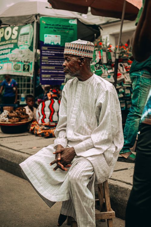 Free Elderly man wearing traditional attire sits calmly amidst a vibrant African market scene. Stock Photo