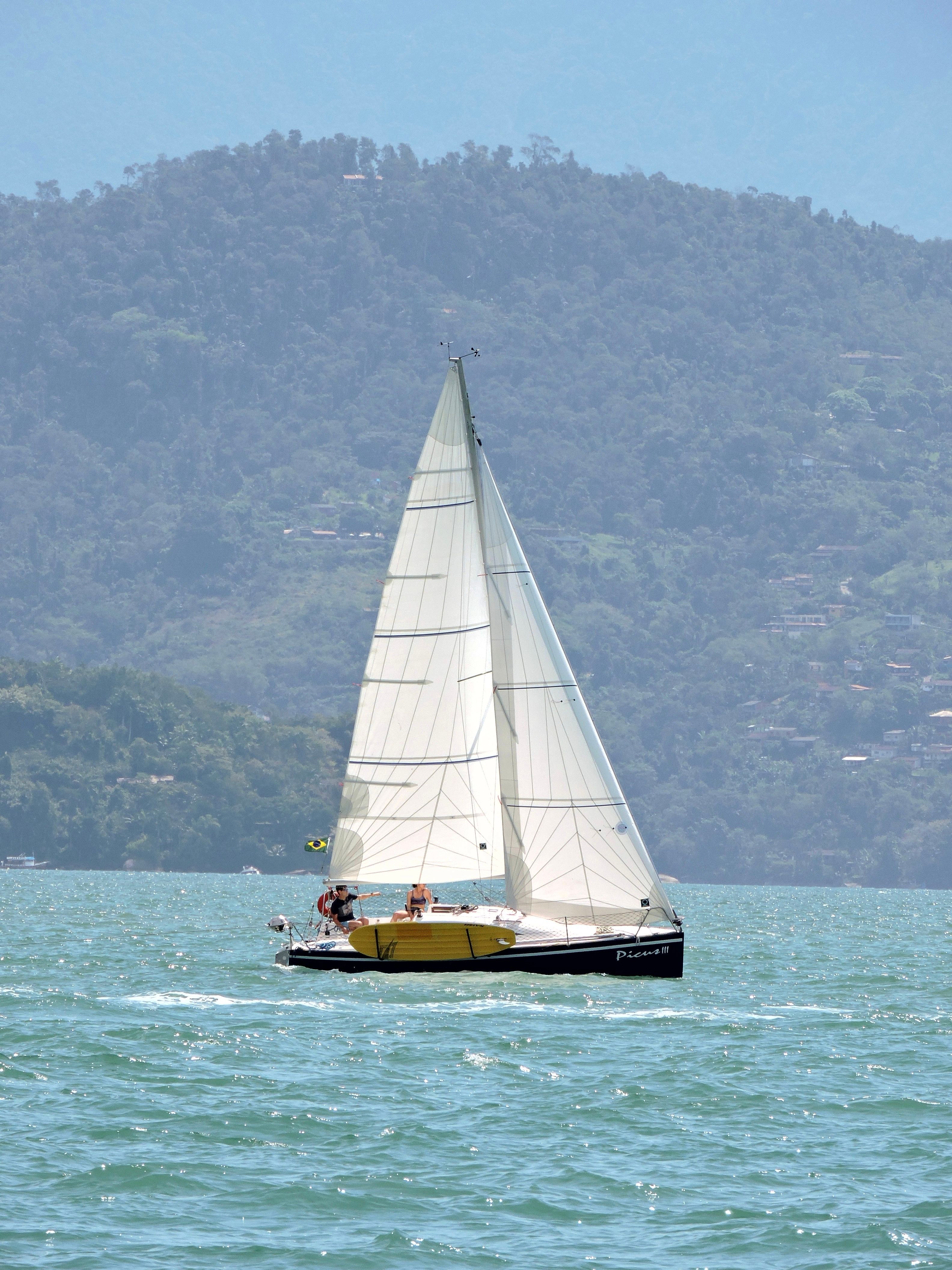 A sailboat with white sails navigates a turquoise sea against a scenic mountain backdrop.