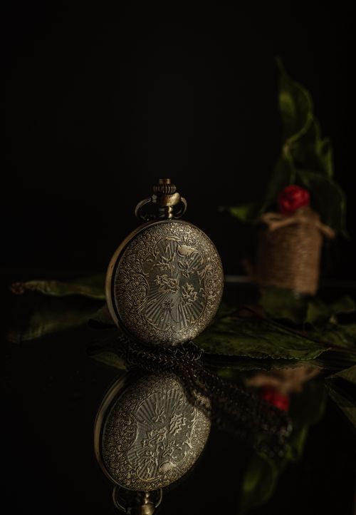Free Intricate vintage pocket watch with floral engravings on a reflective surface, surrounded by leaves. Stock Photo