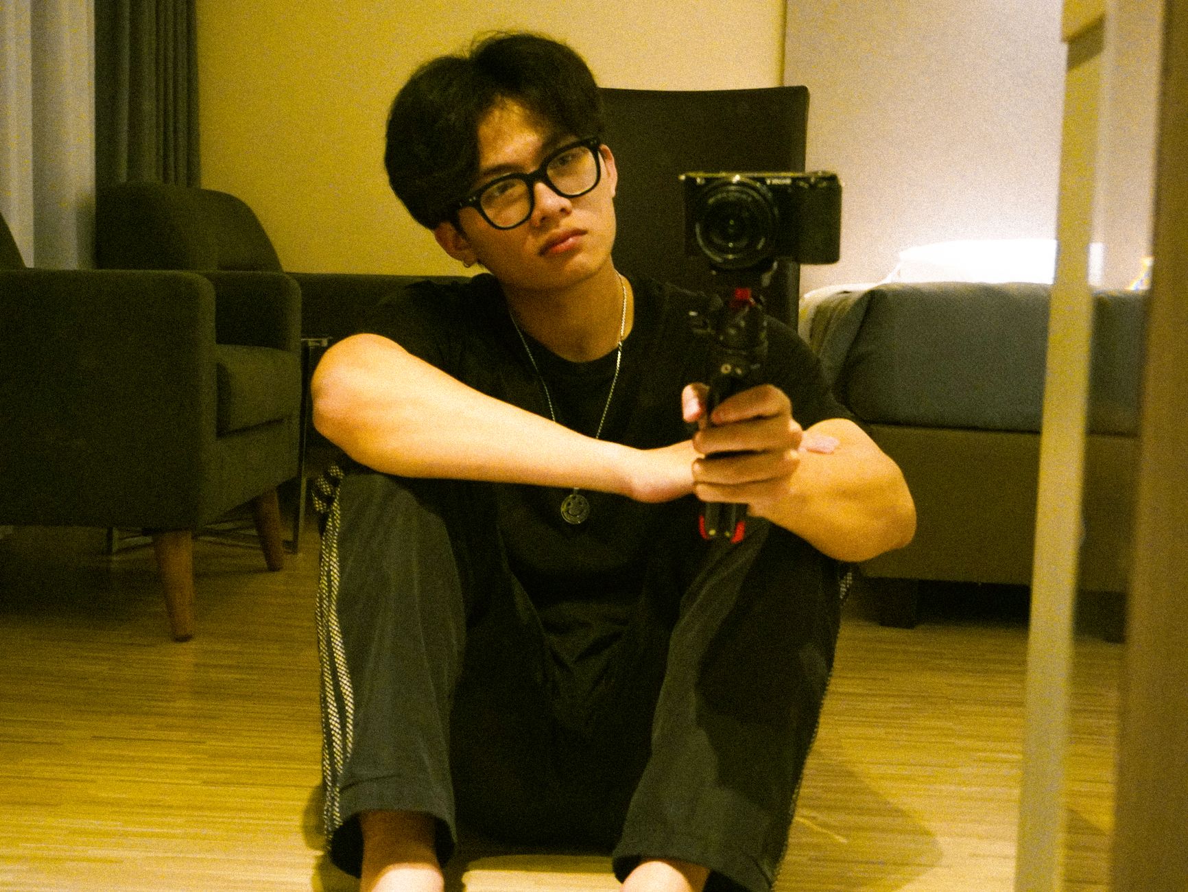 A young man sitting on the floor takes a mirror selfie with a camera, capturing a relaxed indoor setting.